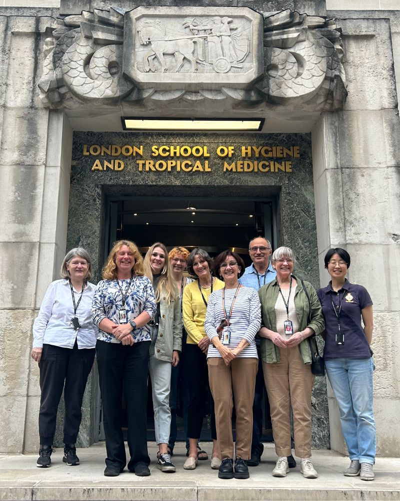 The CureME team are gathered at the top of the stairs at the entrance to the London School of Hygiene and Tropical Medicine. All members smile at the camera. The building is grey with the iconic chariot emblem above them.